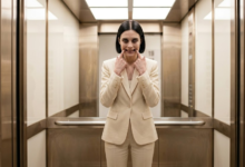 Moteris stovi biuro lifte ir abiem rankų smiliais per jėgą tempdama savo lūpų kampučius į viršų, formuoja nenatūraliai plačią šypseną.