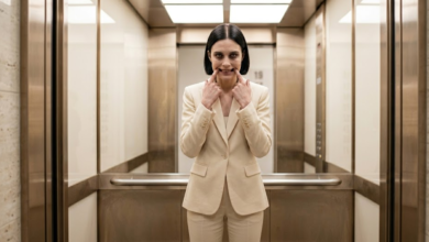 Moteris stovi biuro lifte ir abiem rankų smiliais per jėgą tempdama savo lūpų kampučius į viršų, formuoja nenatūraliai plačią šypseną.