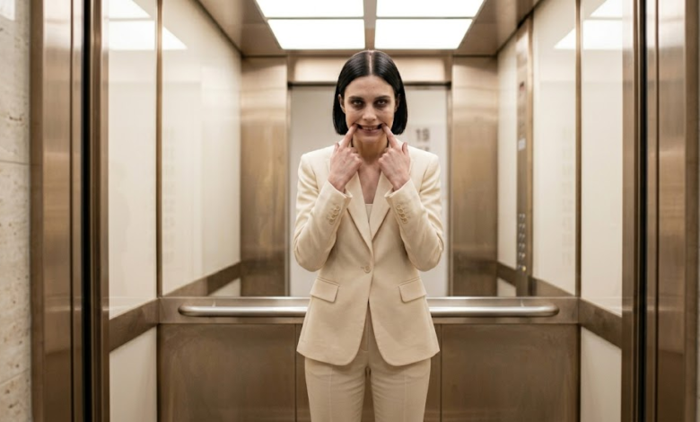 Moteris stovi biuro lifte ir abiem rankų smiliais per jėgą tempdama savo lūpų kampučius į viršų, formuoja nenatūraliai plačią šypseną.