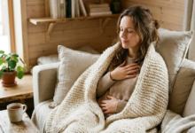 Moteris jaukiai susisupusi į minkštą, stambaus mezgimo pledą, užmerktomis akimis ir lengvai priglaudusi ranką prie krūtinės ar pilvo