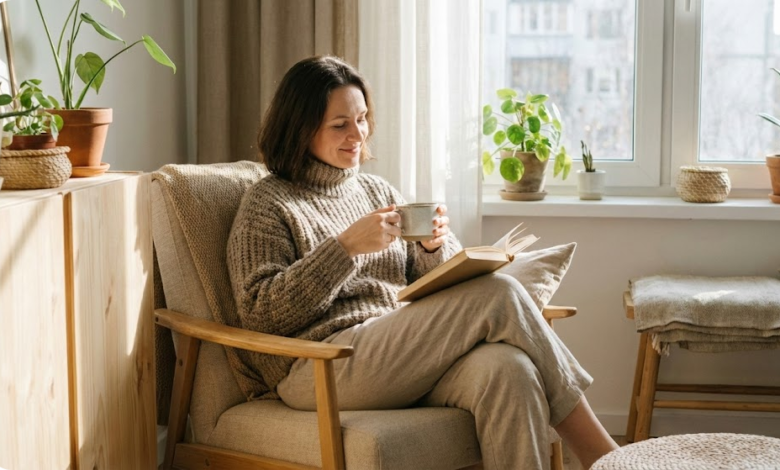 40 metų moteris sėdi fotelyje namie su knyga ir arbatos puodeliu